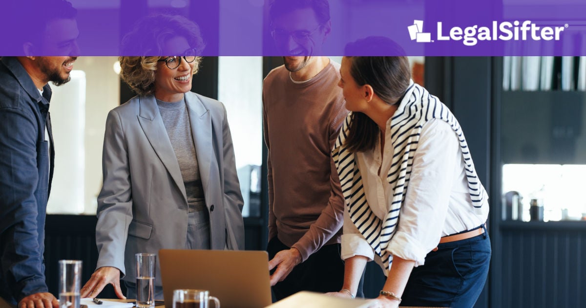 Four colleagues are gathered around a wooden table in a modern office, engaged in a friendly discussion. They are smiling and looking at a laptop screen while collaborating. Two glasses of water and a coffee mug sit on the table, and the atmosphere appears positive and professional.